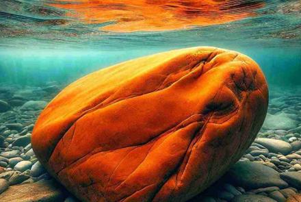 A massive creek rock partially submerged in a flowing stream, surrounded by rippling water.